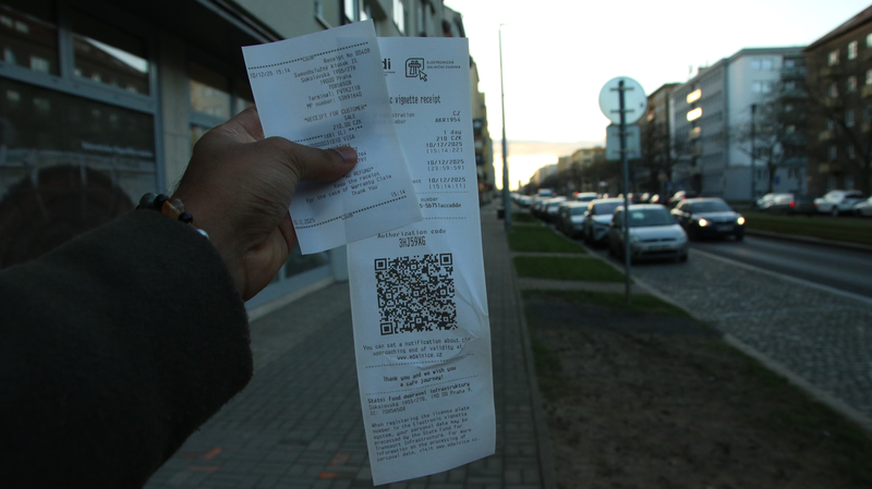 Hand holding a Czech electronic vignette receipt for a 1-day vignette at 210 CZK with QR code and authorization code, purchased from an SFDI self-service machine in Prague on December 10, 2025